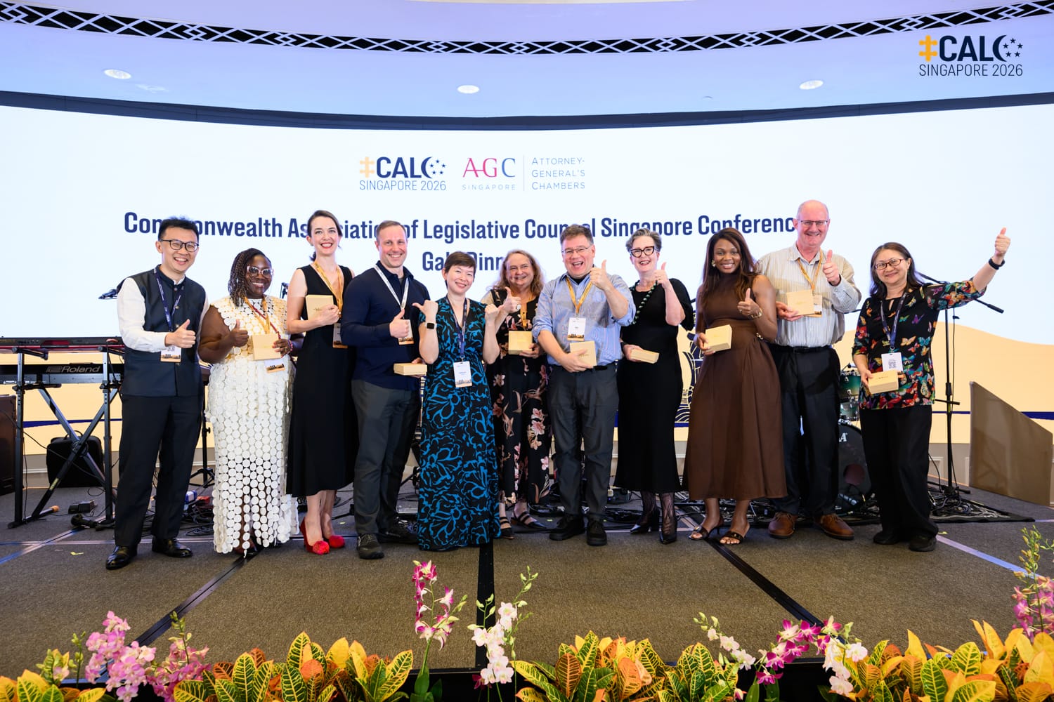 A group of delegates and Ms Jeanne Lee, Chief Legislative Counsel of Singapore, pose on stage with thumbs up at the Conference Gala Dinner, with the event backdrop displayed behind them.